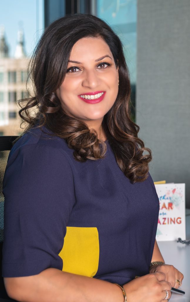 A portrait of Neelam Kaul, standing at a desk and smiling