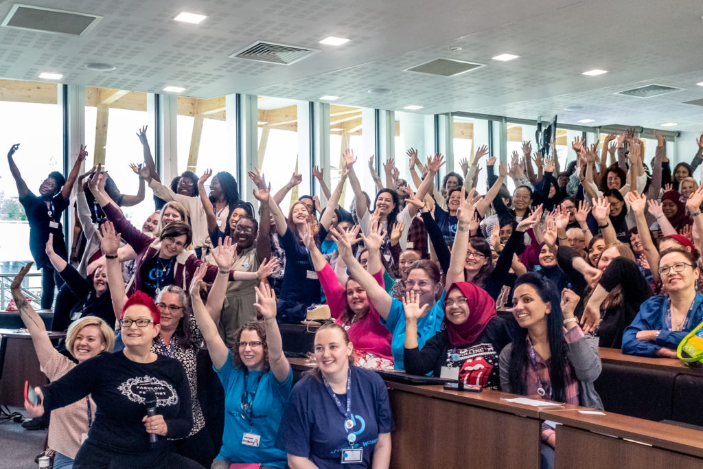 Image shows a large group of diverse women in a lecture theatre with their hands in the air
