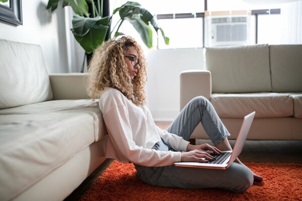 Image shows a woman with blonde curly hair sat on the floor using a laptop.