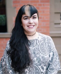 Headshot of Winona Sharpe in front of a red brick wall, Winona has long black hair laid over her shoulder.