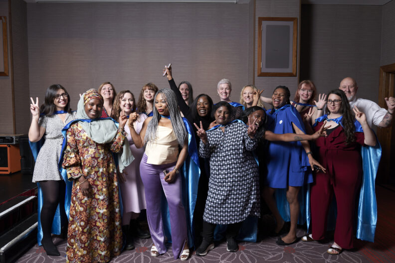 Image shows a varied group of women, some wearing capes, all are smiling and celebrating